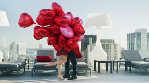 Young beautiful couple having romantic date on rooftop. Two lovers kissing hidden by bunch of colorful red hearts balloons at hotel with city view. Intimacy of Pretty Couple, Modern Love of Young Pair