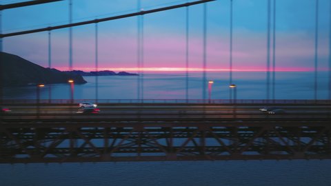 Aerial shot expensive luxury white sport car SUV driving by clean road highway by suspension bridge with cinematic beautiful pink sunset on cloudy day over deep blue ocean. EV cars future concept 4K