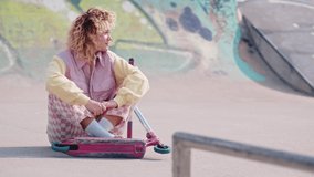 Caucasian teenage girl in retro style clothing sitting at skate park with pink scooter, waving at camera. Slow motion