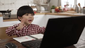 Asian young kid student waving hand to computer screen and talking to his teacher through video conferencing technology while learning from home