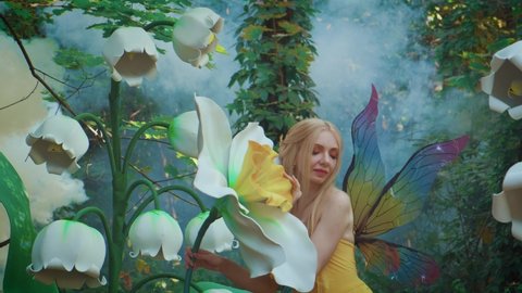 Portrait fantasy woman in fairy costume, bright multi-colored butterfly wings. Elf girl model posing near large artificial narcissus flower, lilies of valley. Background Mystical summer forest in fog