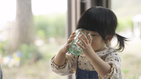 portrait of a thirsty naughty asian little girl drooling and spitting drinking water from cup in restaurant over blurred glass window background.