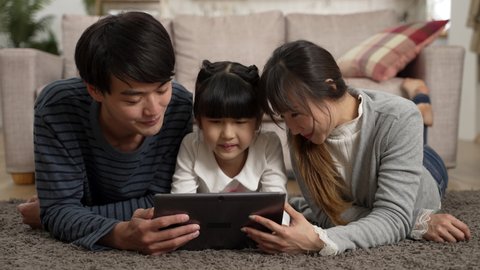 happy Asian family of three watching funny online video with tablet computer together at home. they lie prone on living room floor and point at screen while laughing