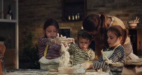 Cinematic authentic shot of happy mother and her little kids are having fun to make dough with flour and knead it together for baking cookies in kitchen at home.