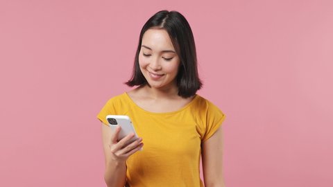 Excited joyful young woman of Asian ethnicity 20s wears yellow t-shirt using mobile cell phone hold fan of cash money in dollar banknotes isolated on plain pastel light pink background studio portrait