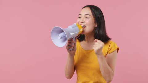 Fun promoter young woman of Asian ethnicity 20s wears yellow t-shirt point index finger aside scream in megaphone announces sale Hurry up isolated on plain pastel light pink background studio portrait