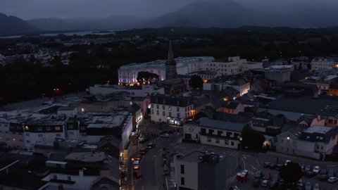 Aerial footage of town centre with church and luxury hotel at dusk. Backwards reveal of buildings along illuminated street. Killarney, Ireland