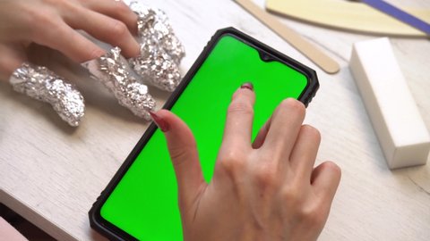 A girl during a manicure uses a smartphone with a green screen. Selfmade manicure service. Manicurist paints nails with pink gel polish. Manicured red nails. Nail polish application.