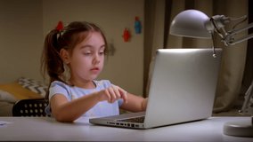 Preschool girl studying online using a laptop. A girl child is watching an Internet video course while sitting at a writing desk at home.