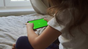 View over the shoulder of a Preschool Girl At Home Holding a Smartphone with a Chrome Green Screen And Viewing Content. Child, Using A Mobile Phone, Browsing Internet Content, Video