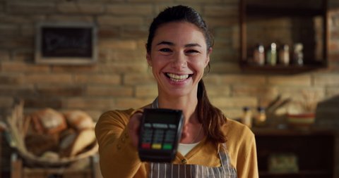 Cinematic shot of young friendly smiling saleswoman showing in camera atm terminal for contactless payments and NFC technology service in bakery shop.