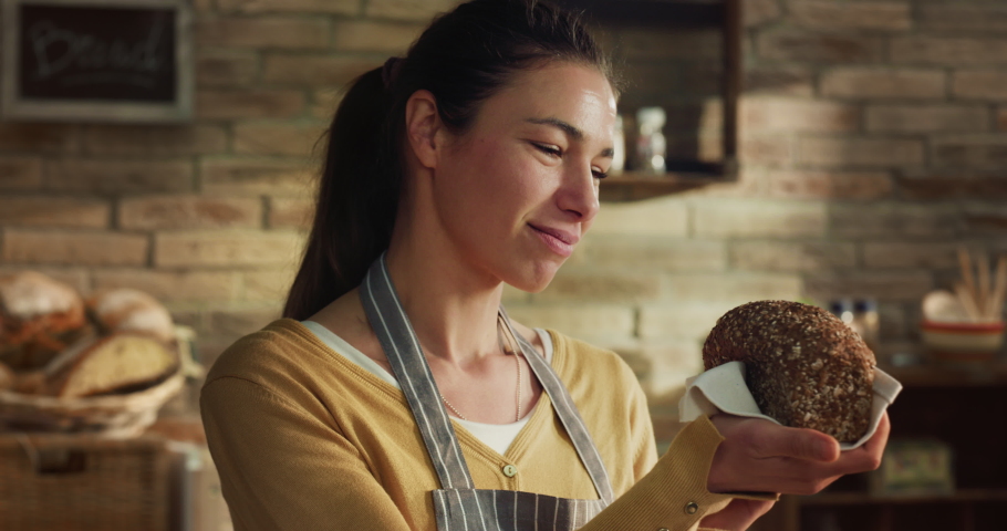 Cinematic close up shot of young female artisan baker is smelling fresh baked bread taken out of oven at moment to control quality and smiling satisfied in rustic bakery shop kitchen. Royalty-Free Stock Footage #1088744979