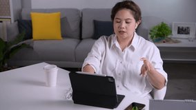 Happy Asian woman wearing earphones having a video call on a tablet sitting on a desk in the living room at home. Young Asian woman entrepreneur giving online educational class lecture