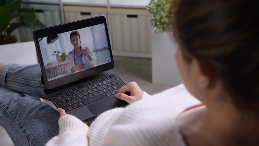 Young Asian woman sit on couch at home have video call with doctor use wireless internet connection on laptop, female patient talk consult with physician online on webcam conference on computer