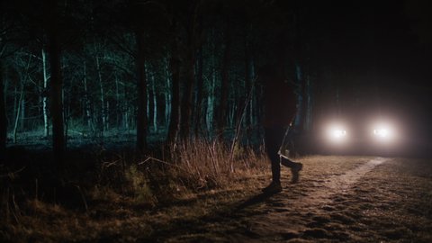 Car headlights in an empty forest at night, silhouette of a mysterious man entering dark woods alone. Thriller scene.
