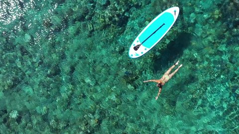 The girl swims in the crystal clear sea next to a paddleboard. Woman with SUP board in turquoise sea. Aerial top down shot