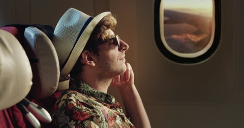 Cinematic shot of young man wearing straw hat and sunglasses sitting in cabin of aircraft and enjoying traveling with comfort during international flight to summer holidays vacation trip.