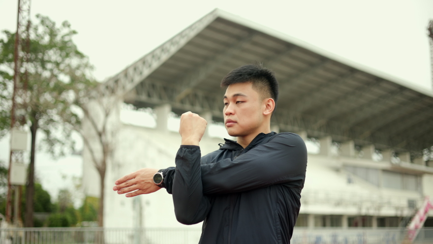 Young asian man wearing sportswear warming up front of sport stadium. Chinese man stretching arms before running on track exercise in the morning. Healthy and athlete lifestyle concept.