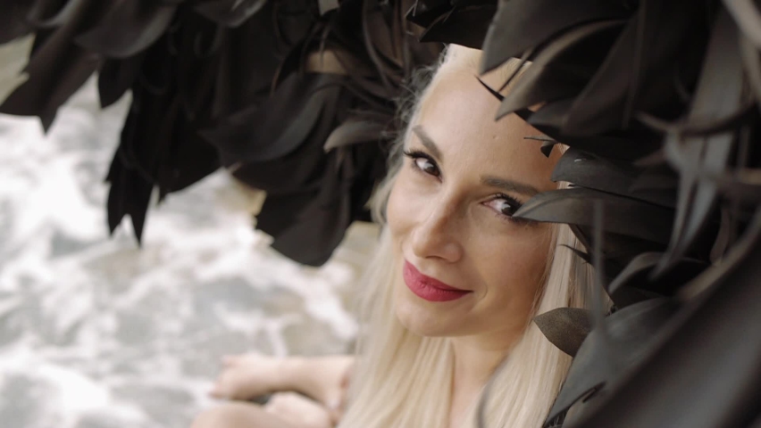 Blond young woman dressed as angel with black wings. Fashion sexy female model on sea beach looking at camera. High angle view. Close-up.