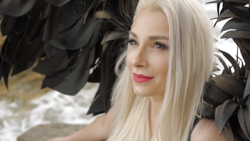 Sexy blond young woman dressed as angel with black wings. Fashion female model looks up sitting on the stone above the sea. Waves are splashing below. Close-up.