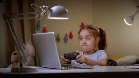 Cheerful preschool girl, playing video games with a joystick late at night on a laptop with a controller in her hands - the concept of children's gaming addiction and late-night games