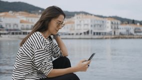 A pretty girl, a brunette, of Asian appearance, sits on a pier, on the seashore, against the backdrop of a small town, smiles, scrolls her phone and looks into the distance. Medium plan.