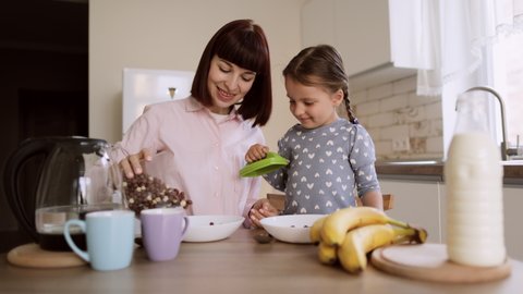 Breakfast in the Kitchen. Young Beautiful Mother Pours cornflakes into Bowl. Adorable Little Daughter Preparing to Eat with Pleasure. Caring Mother Prepares cornflakes for Her Cute Little Girl.