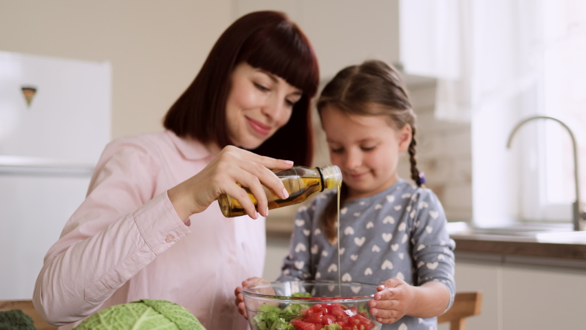 Portrait of smiling mom and little daughter adding olive oil to the salad while cooking in kitchen together, happy mother and small girl child have fun preparing healthy food at home Royalty-Free Stock Footage #1088753821