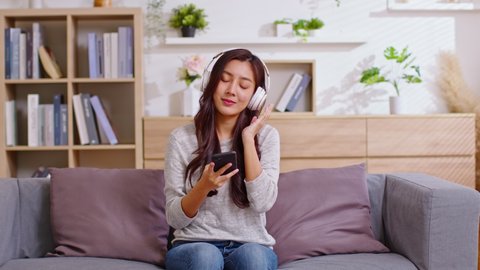 Young women listening to music in headphones and dancing energetic and rhythm at couch. Attractive Asian girl looking at the smart phone sitting in the living room spending leisure time at home.