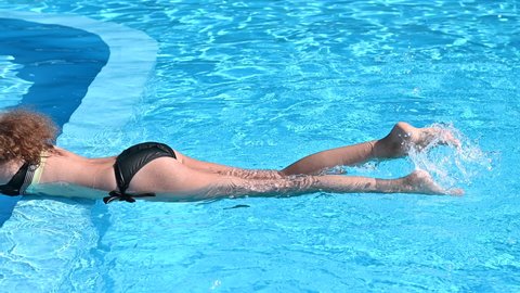 Relaxed girl resting at summer resort on sunny day. Side view of slim female tourist liying on water of pool and splashing water. 