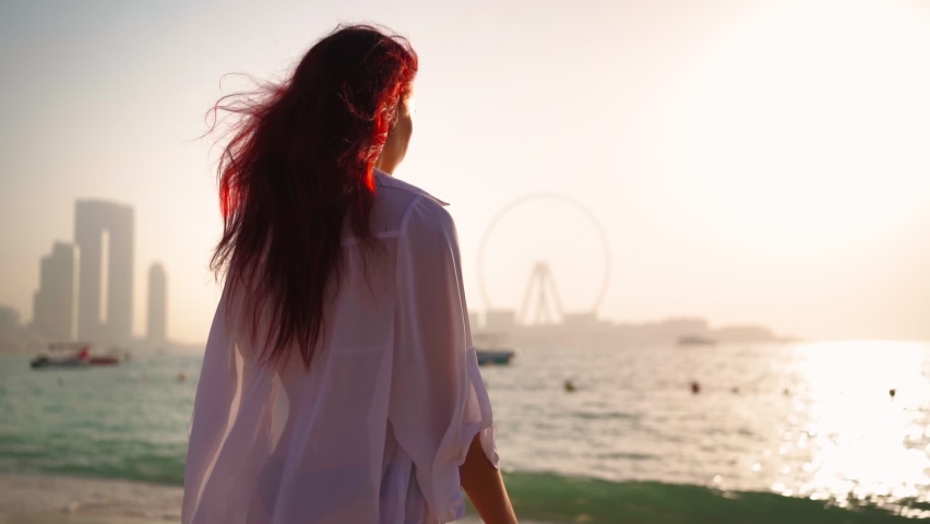 Woman in a white shirt walks along the beach of the Persian Gulf in Dubai towards the sea against the backdrop of a beautiful sunset. Journey through the United Arab Emirates. Slow motion. 