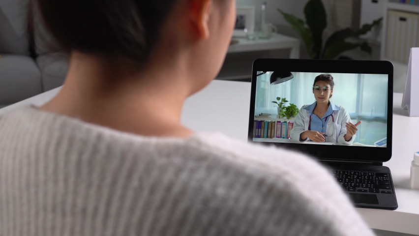 Young Asian woman sit on couch at home have video call with doctor use wireless internet connection on laptop, female patient talk consult with physician online on webcam conference on computer