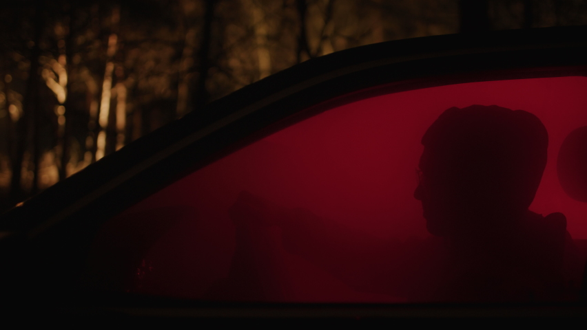 Creepy man sitting in his car at night in the forest, red light. Silhouette of a dangerous stalker isolated in the woods at night. | Shutterstock HD Video #1088760411