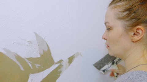 a woman removes old wallpaper from the wall and prepares to make repairs in the room