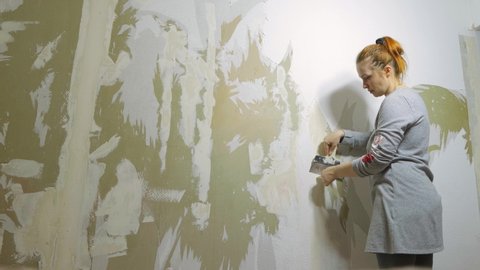 a woman removes old wallpaper from the wall and prepares to make repairs in the room