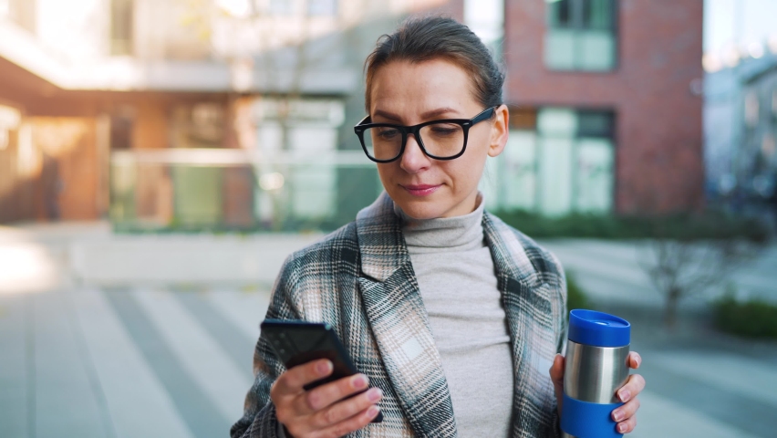Caucasian businesswoman with glasses and a coat walks through the business district, with thermo cup and using smartphone. Communication, work day, busy life concept