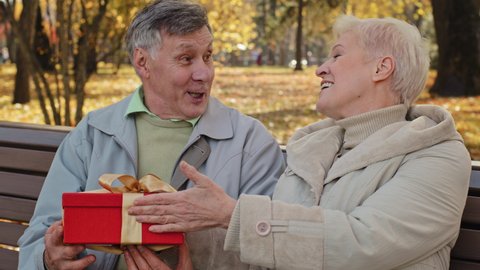Happy smiling elderly couple sitting on bench in autumn park mature old grandma give gift husband make surprise excited joyful grandfather kissing wife on cheek celebrate wedding anniversary birthday