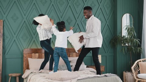 Happy african family parents with daughter standing on bed hitting pillows having fun at home in bedroom father and mother playing with little girl child kid funny fight morning game laughing together