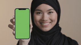 Portrait asian girl muslim islamic woman in black hijab smiling showing mobile phone with green screen holding smartphone looking at camera promo video gadget advertisement device telephone online