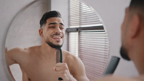Close-up view from behind male reflection in mirror naked arab guy millennial bearded handsome cheerful happy man sings in hairbrush having fun singing song dances to music in bathroom shower morning