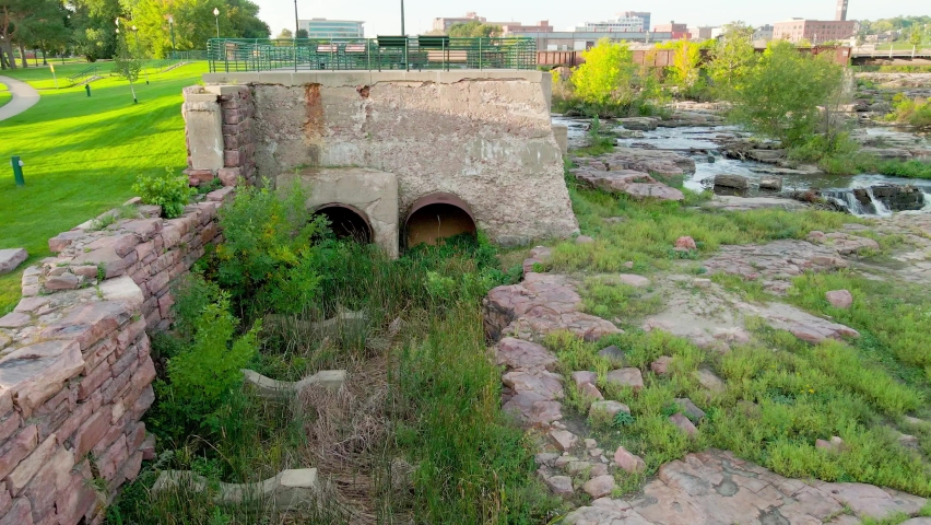 Pan out from observation point from Falls Park in South Dakota. Ruins from an old mill preserved in this park. Sunlight gleaming on the grass through the shadows. Water flowing over the stones. 
