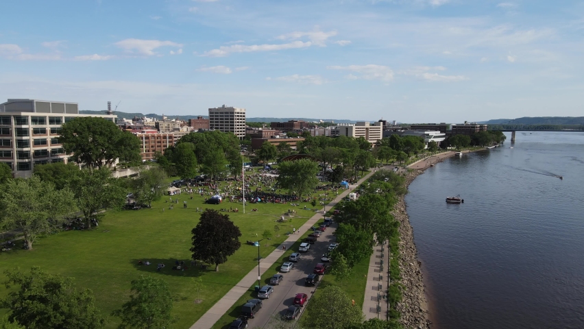 Flyover Riverside Park, La Crosse, Wisconsin, along the Mississippi River during music night in the park on a summer day. Downtown office buildings. Bluffs with forest covering. 