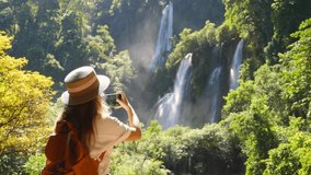 Back view of millennial travel woman on trip or adventure. Young travel blogger on waterfall, make photos on smartphone for social media. Amazing landscape of Thi Lor Su waterfall in Thailand