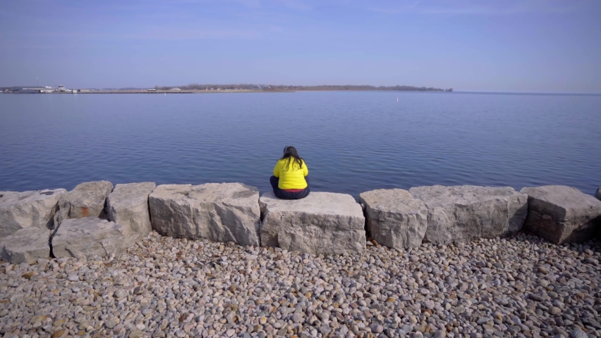 Person in Toronto Trillium Park with headphones sitting near lake in bright yellow jacket during day. Provincial park developed along Lake Ontario. ON, Canada