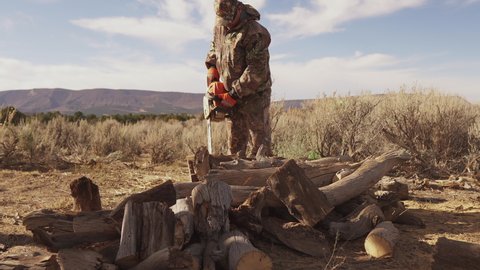 A man in hunting gear uses a chainsaw to cut up firewood. Camo gear.