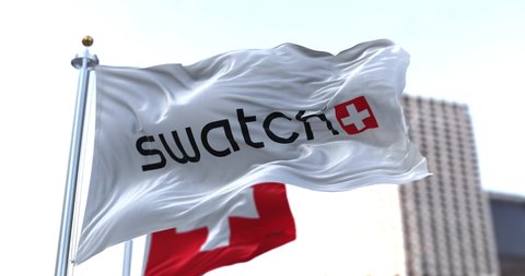 Bern, CH, March 2022: the flag with the Swatch logo waving in the wind with the national flag of Switzerland blurred in the background. Swatch is a Swiss watch company