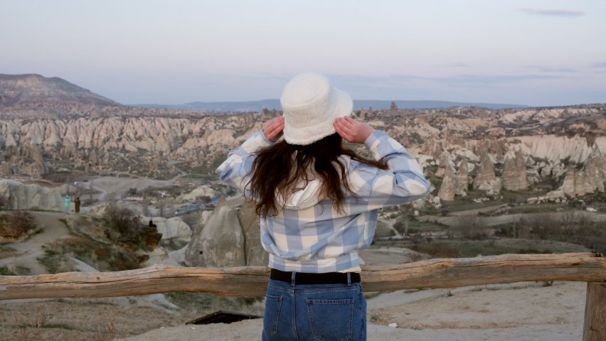 Woman in checkered shirt holding white hat and look at stone valley in Cappadocia Turkey, enjoy beautiful view. Back View Scenery