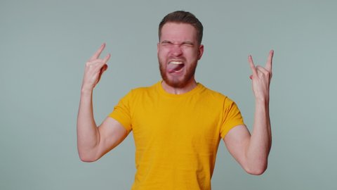 Rock n roll! Overjoyed delighted man 20 years old in t-shirt showing gesture by hands, cool sign, shouting yeah with crazy expression, dancing, emotionally rejoicing in success win alone on gray wall