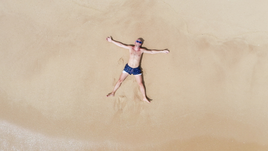 4K drone slowly rising above man lying in star-shaped pose on sandy beach. Summer holiday, ocean holiday, relaxation concept. Royalty-Free Stock Footage #1088779747