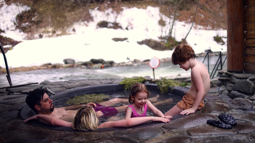 happy family with kids relaxing in hot bath tub outdoors, enjoying thermal spa in stone vat on fire. winter activity and leisure